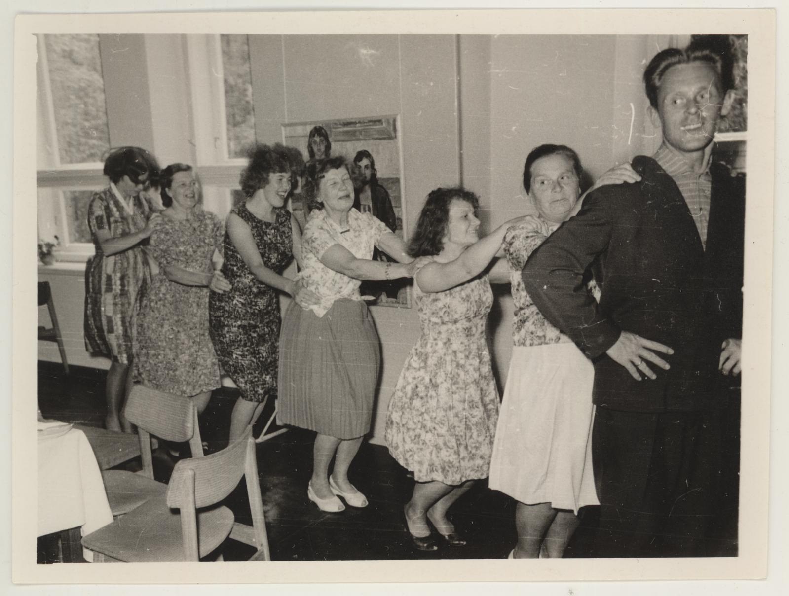 Eine Gruppe von Menschen, die in einem Raum mit Stühlen und einem mit wei├čer Decke bedeckten Tisch tanzen. An der Wand befindet sich ein Photo-Rahmen und im Hintergrund sind Fenster zu sehen. Die Menschen in dem Bild tragen Kleidung aus den 1950er Jahren.