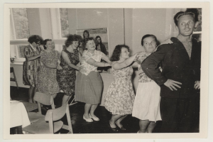 Eine Gruppe von Menschen, die in einem Raum mit Stühlen und einem mit wei├čer Decke bedeckten Tisch tanzen. An der Wand befindet sich ein Photo-Rahmen und im Hintergrund sind Fenster zu sehen. Die Menschen in dem Bild tragen Kleidung aus den 1950er Jahren.