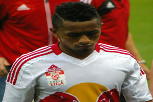 Ein Mann in rot-weißem T-Shirt steht auf einem Fußballfeld und schaut entschlossen, mit zwei weiteren Personen in rotem T-Shirt im Hintergrund.