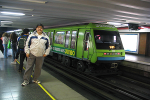 Ein Zug auf Schienen mit Menschen, die links stehen und ein Dach oben im Bild.