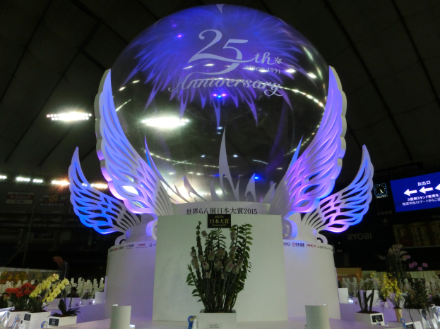 Ein großer Kristallball mit Flügeln oben drauf, umgeben von Blumenbouquets, Namensschildern und anderen Gegenständen auf dem Boden, mit Schildern, Lichtern und einem Bildschirm im Hintergrund bei einer festlichen 25-jährigen Jubiläumsfeier.