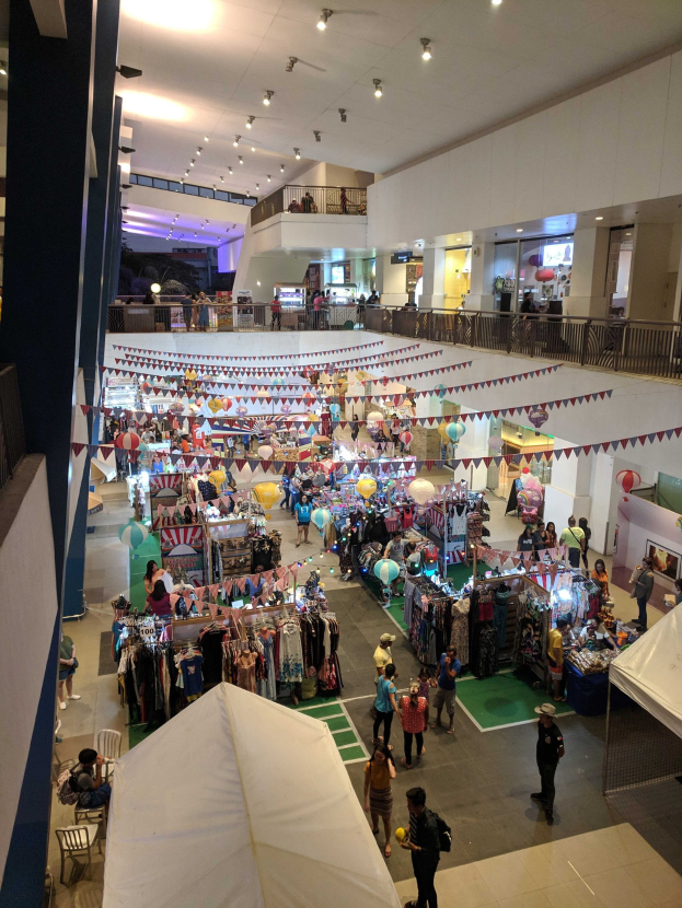 Ein volles Einkaufszentrum mit Kleiderständen, Dekorationsfahnen, Ballons, sitzenden und stehenden Menschen, Geländern, Säulen und Deckenbeleuchtung, das an einen Weihnachtsmarkt im Mall of Asia erinnert.
