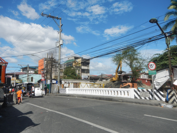 Eine belebte Stadtstraße mit Fahrzeugen, Fußgängern, Strommasten, Schildern, Bäumen, Gebäuden und einem bewölkten Himmel, mit einer Baustelle in der Mitte der Straße.