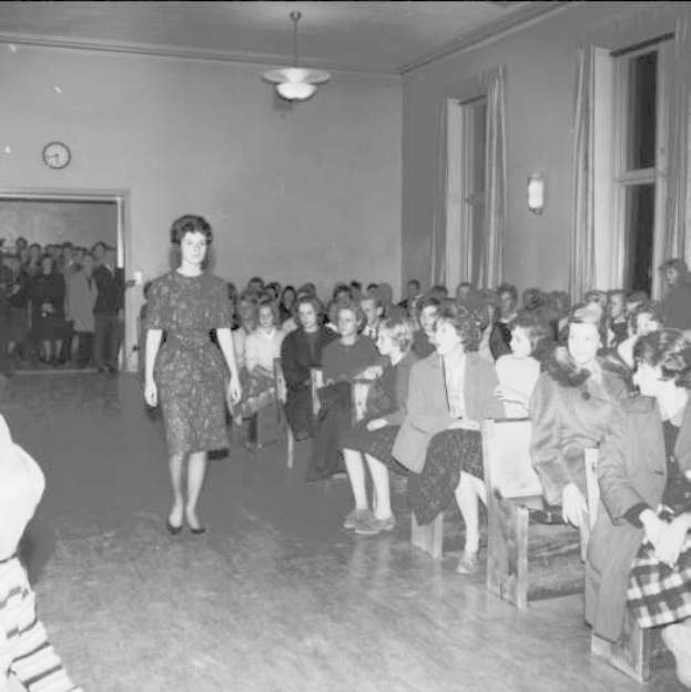 Eine Frau in einem schwarzen und weißen Kleid läuft über einen Laufsteg während einer Modenschau für Frauenrechte, mit einer Menge, die auf Stühlen sitzt und einer Uhr und Lampen an der umliegenden Wand.