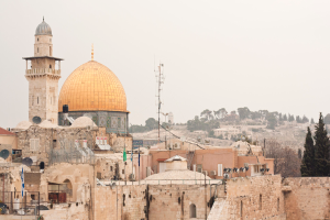 Mehrere Backsteingebäude mit Satellitenschüsseln und Klimaanlagen vor Wald und Himmel.