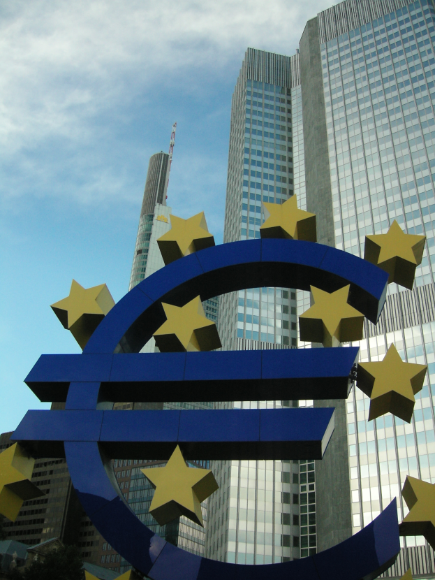 Das Euro-Zeichen steht prominent vor dem Hauptquartier der Europäischen Zentralbank, einem großen modernen Gebäude, das von Bäumen umgeben ist und unter einem bewölkten Himmel liegt.