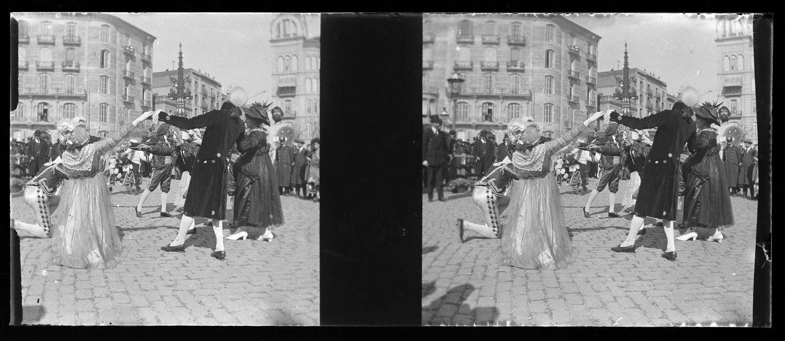 Schwarzes und weißes Foto von Menschen in Kostümen, die auf einer Straßenparade tanzen, umgeben von Gebäuden, Straßenlaternen und Lichtern unter einem klaren Himmel.