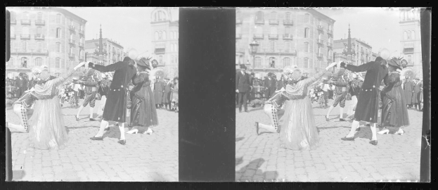Schwarzes und weißes Foto von Menschen in Kostümen, die auf einer Straßenparade tanzen, umgeben von Gebäuden, Straßenlaternen und Lichtern unter einem klaren Himmel.