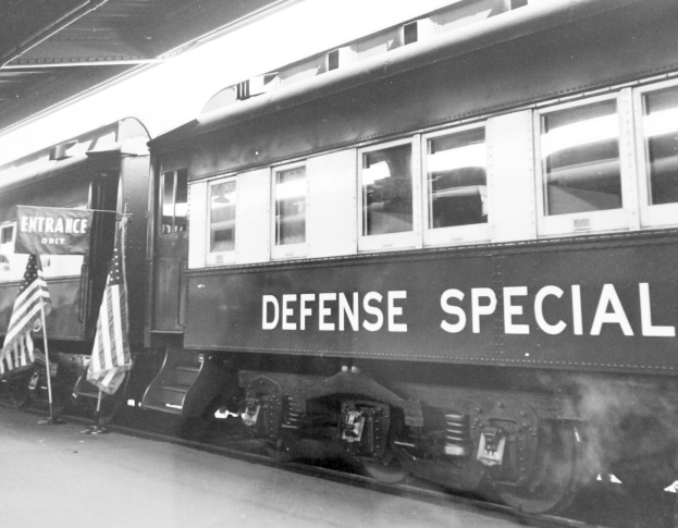 Schwarz-weiß-Foto eines Zuges mit der Aufschrift "Defense Special" auf den Schienen mit zwei Fahnen links und einer Decke darüber.
