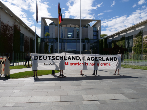 Gruppe von Menschen mit einer Fahne, auf der "Deutschland, Lagerland Migration ist kein Verbrechen" steht, vor einem Gebäude mit Fenstern, umgeben von Gras, einem Metallzaun, Pflanzen, Bäumen und Fahnen auf Stangen unter einem bewölkten Himmel.