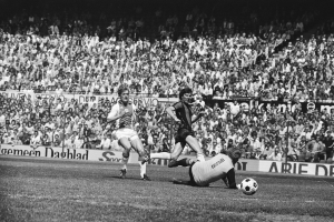 Schwarzes und weißes Foto von Männern, die auf einem Feld Fußball spielen, mit Zuschauern im Hintergrund und einer Fahne mit der Aufschrift "Bundesliga 1978-79 Ajax 1-1 Hannover 96".