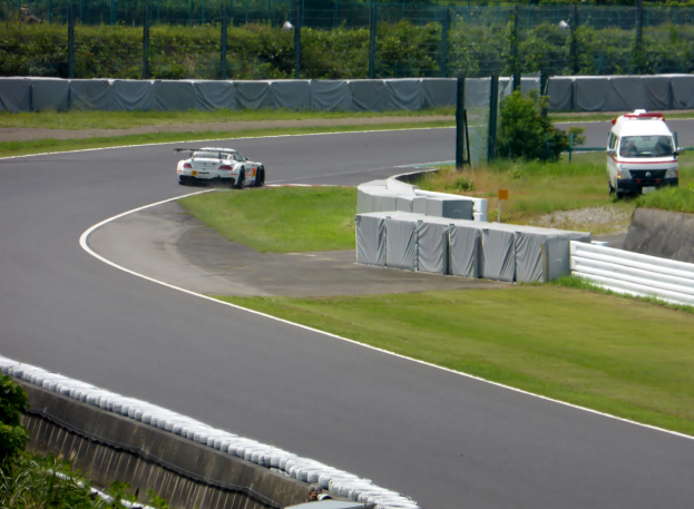 Ein Rennwagen fährt auf einer Rennstrecke mit einem Polizeiwagen im Hintergrund, umgeben von Vegetation und Absperrungen, mit Text am unteren Bildrand.