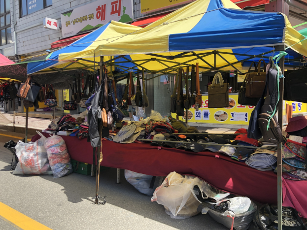 Ein Straßenmarkt mit Zelten, die Taschen, Hüte und andere Gegenstände präsentieren, vor Häusern mit Fenstern und Texttafeln.