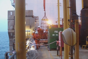 Ein Schiff mit Masten, Kisten und Geländern, das Menschen mit Helmen zeigt, vor einem Wasserhintergrund.