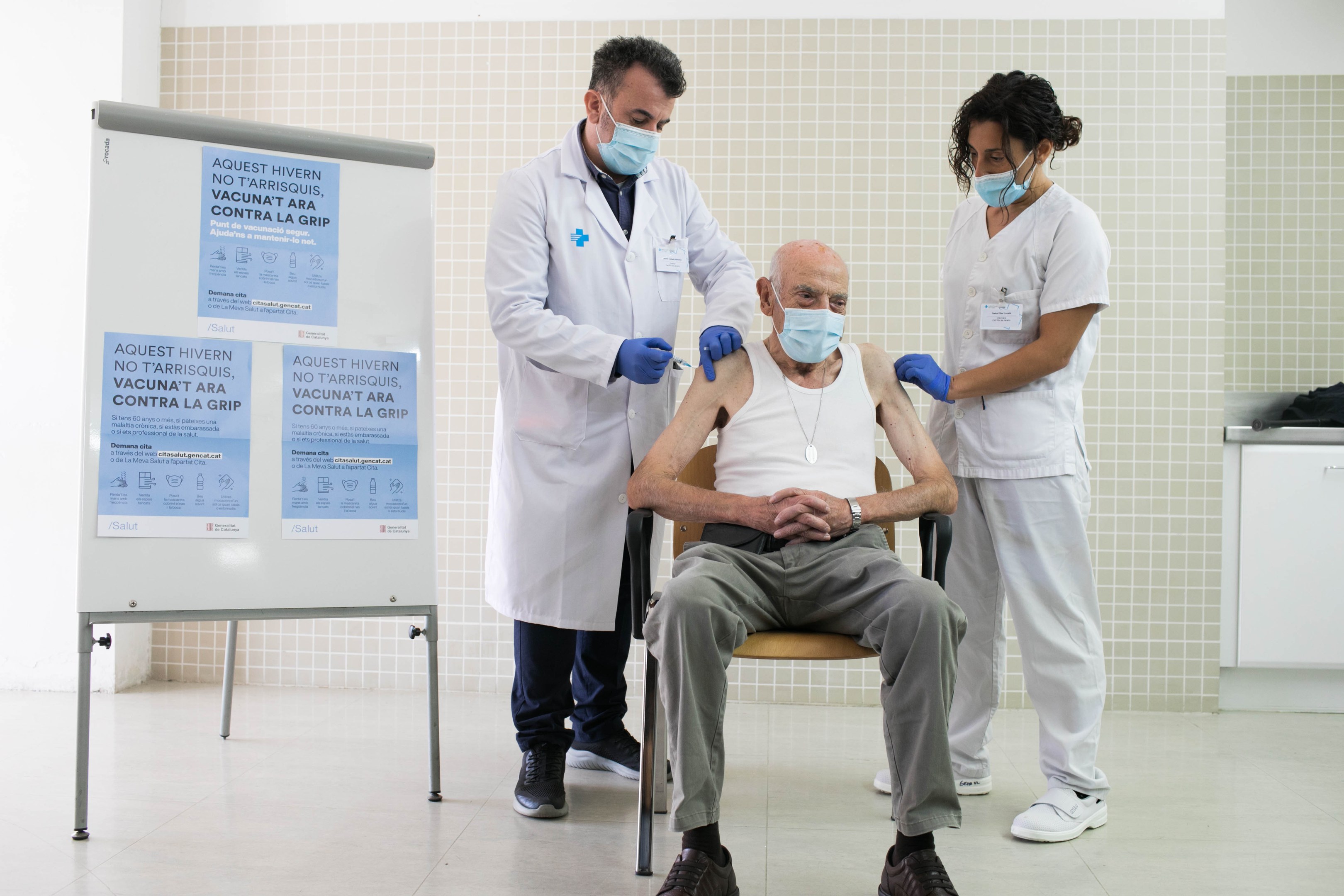 Alter Mann sitzt in einem Stuhl und erhält eine Impfung von zwei maskierten Ärzten in einem Krankenhauszimmer, mit einer Tafel und medizinischen Gegenständen in der Nähe.