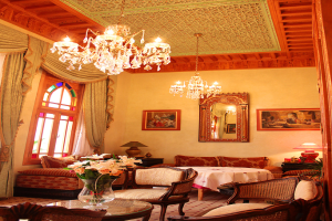 Ein Wohnzimmer mit einem Tisch und Blumentopf im Vordergrund, umgeben von Stühlen, Sofas und einem Tisch mit einer wei√üen Decke im Hintergrund, gerahmte Bilder an der Wand, von der Decke h√§ngende Lichter und Vorh√§nge mit einem Fenster links.