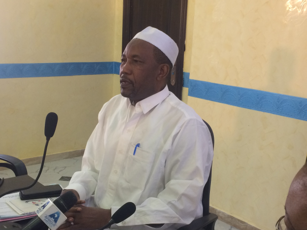 Ein Mann in einem weißen Gewand und einer Kappe sitzt an einem Tisch mit Mikrofonen, spricht, während Papiere und Gegenstände auf dem Tisch zu sehen sind, mit einer anderen Person mit Brille zu seiner Rechten, in einem Raum mit einer Wand und einer Tür dahinter.