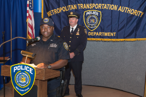 Zwei Polizeibeamte stehen vor einem Rednerpult, einer hält Papiere und spricht in ein Mikrofon, mit einer Fahne, einem Tisch und einer Tafel mit Symbolen und Text dahinter.