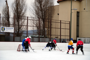 Menschen beim Eis-hockey-Spielen auf einer Eisfläche mit Gebäuden, Bäumen, einer Straßenlaterne, einem Namensschild und Zäunen im Hintergrund vor einem Himmel.