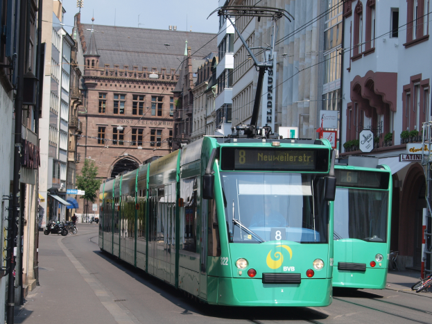 Zwei grüne Straßenbahnen fahren eine von Hochhäusern gesäumte Stadtstraße entlang, mit parkenden Fahrrädern und Fußgängern auf den Gehwegen, unter einem klaren blauen Himmel mit einem Baum im Hintergrund.