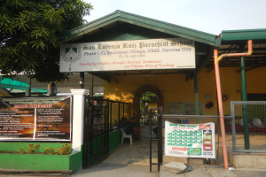 Der Eingang der San Lorenzo Ruiz Pfarrschule, mit einer Tafel mit Text, einem Metallzaun, Menschen, die unter einem Dach sitzen, Pfosten, einer Schautafel, Pflanzen, einem Baum und einem bewölkten Himmel.