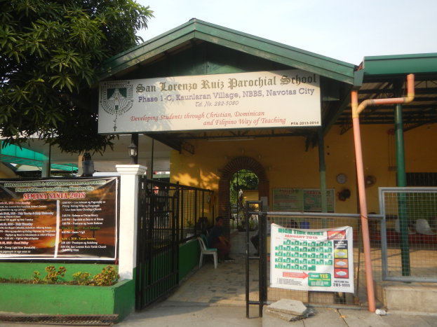 Der Eingang der San Lorenzo Ruiz Pfarrschule, mit einer Tafel mit Text, einem Metallzaun, Menschen, die unter einem Dach sitzen, Pfosten, einer Schautafel, Pflanzen, einem Baum und einem bewölkten Himmel.