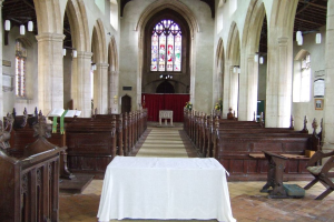 Ein Raum mit Tischen, einem Podium, einem Bücherregal, Stühlen, Bänken, Säulen, Brettern, einem Vorhang, Fenstern, Stoff, Pflanzen und verschiedenen Gegenständen.