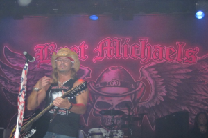 Eine Musikkonzertszene mit Scheinwerfern oben, einem Banner im Hintergrund, Drogen in der Mitte unten, einer Person in der rechten unteren Ecke sitzend und einem Gitarristen mit Mikrofon links.