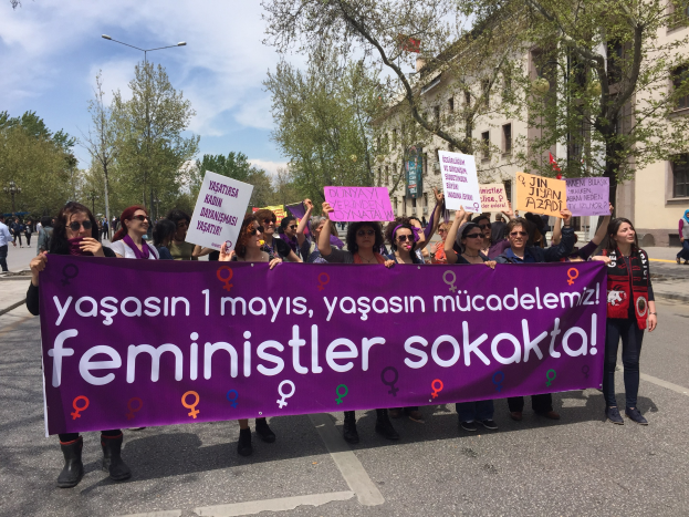 Eine Gruppe von Menschen geht eine Straße entlang und hält ein lilafarbenes Banner mit der Aufschrift 'feminist sokokta' hoch, mit Bäumen und Laternenmasten an der Straße und einem Gebäude im Hintergrund unter einem bewölkten Himmel.