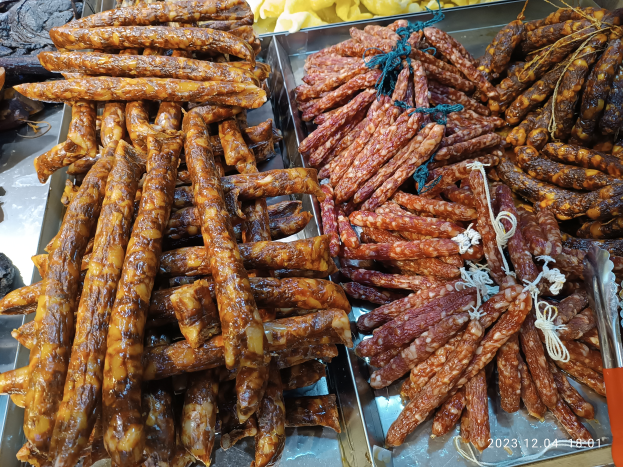 Ein Tisch mit verschiedenen Würstchensorten in Schalen, umgeben von anderen Speisen und einer Person im Hintergrund auf einem Markt.