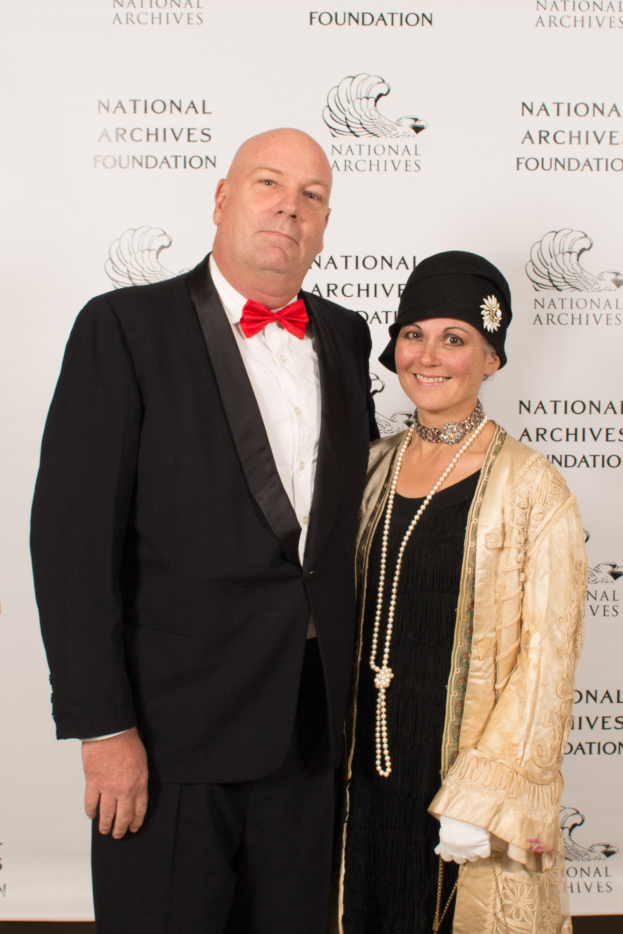 Ein Mann in einem schwarzen Anzug mit einem weißen Hemd und schwarzer Fliege steht neben einer Frau in einem eleganten Kleid mit einem gemusterten Schultertuch auf einem roten Teppich, mit einem weißen Banner mit Text im Hintergrund.