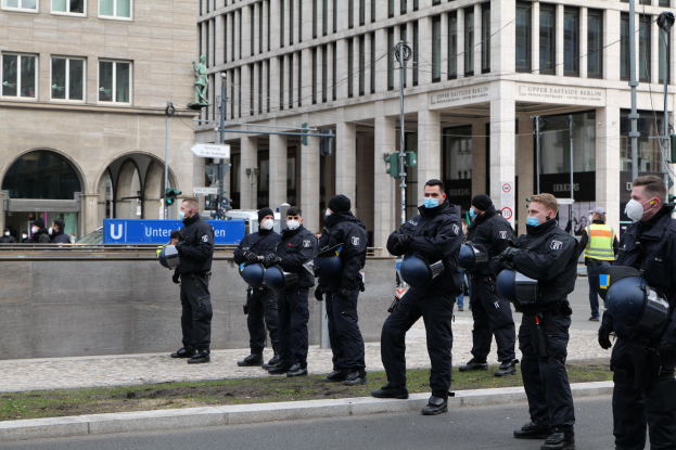 Eine Gruppe von Polizisten in schwarzen Uniformen und Masken steht vor einem gläsernen Gebäude mit Säulen in Berlin, Deutschland, einige halten Helme, mit Laternen, Verkehrszeichen, Texttafeln, einer Statue, anderen Menschen und grasbewachsenem Boden im Hintergrund.