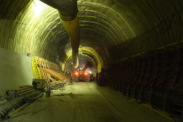 Baustelle mit einem großen Tunnel, Fahrzeugen, verstreuten hölzernen Gegenständen, Rohren, einer Wand auf der linken Seite und beleuchteten Lampen im Hintergrund des Tunnels.
