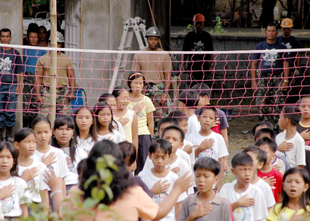 Gruppe von Kindern vor einem Netz mit einer Frau im Vordergrund, Hintergrund enthält Personen mit Helmen, eine Leiter, eine Wand und Bäume.