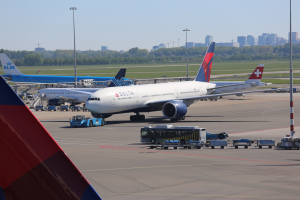 Ein Boeing 777-300ER der Delta Air Lines am Frankfurter Flughafen, umgeben von Fahrzeugen, Laternenmasten, Bäumen, Gebäuden und einem klaren blauen Himmel.