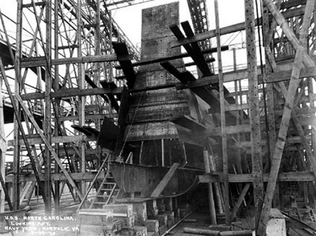 Schwarzes und weißes Foto eines Schiffes in einem Schiffbauhof, mit Holzpfählen und Treppen im Vordergrund und Himmel im Hintergrund; Text "Marinebewaffnung" in der linken unteren Ecke.