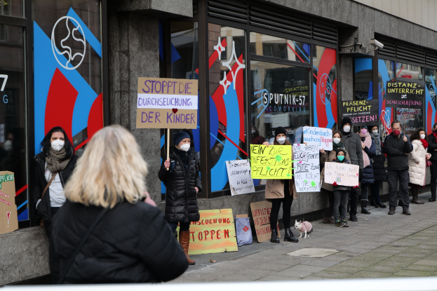 Eine Gruppe von Menschen mit Masken und Schildern protestiert vor einem Gebäude mit Glaswänden und -türen, während Tafeln mit Text und Logos sowie Kameras und ein Hund unten zu sehen sind.