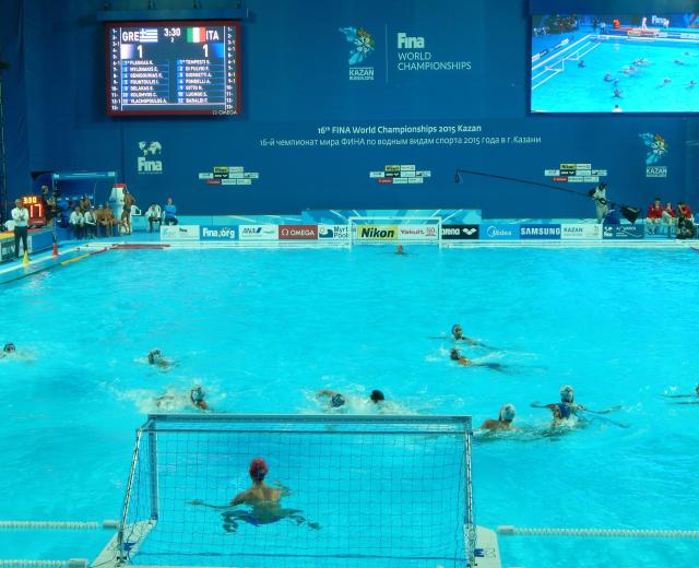 Wasserballspieler bei einem Poolwettkampf während der 2016 FINA-Weltmeisterschaften in Kazan, mit Zuschauern auf der linken Seite und einem Werbeschirm im Hintergrund.