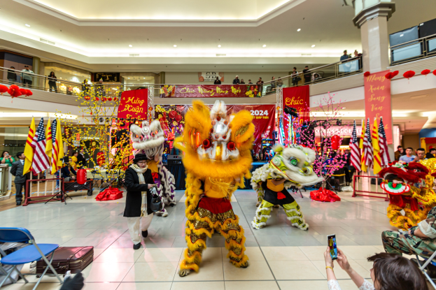 Ein lebendiges chinesisches Neujahrsfest in einem Einkaufszentrum mit Menschen in Kostümen, die tanzen, andere sitzen und verwenden Handys, umgeben von Flaggen, Bannern, Dekorationen, Geländern, Säulen und Deckenlampen.