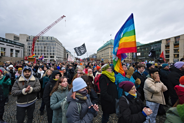 Große Gruppe von Menschen bei einer LGBTQ+-Rechtsdemo in Berlin, die Fahnen und Transparente schwenken, mit Gebäuden, einem Kran und einem bewölkten Himmel im Hintergrund.