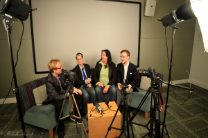 Eine Gruppe von drei Personen sitzt auf einem Sofa und gibt ein Interview, wobei eine Person links in einem schwarzen Mantel und Hose, eine Person in einem Mantel und blauer Krawatte mit einem Mikrofon in der Mitte und eine Person rechts lächelt. Ein Mikrofonrekorder befindet sich auf dem mittleren Tisch, Kameras sind auf der rechten und linken Seite und große Lichtstative sind vorhanden, mit einem Projektionsschirm im Hintergrund.