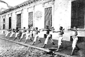 Eine Gruppe von M√§nnern, die Karate auf einem Kopfsteinpflasterstra√üen vor einem Geb√§ude mit Fenstern und T√ľren √ľben.