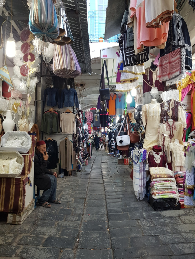Ein belebter Straßenmarkt in der Altstadt von Jerusalem mit Ständen, die Kleidung, Taschen, Lampen und andere Artikel verkaufen, während Menschen im Hintergrund durch die Gassen schlendern und mit einem Gebäude im Hintergrund interagieren.