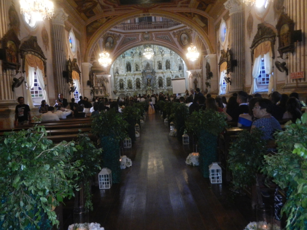 Eine Trauung in einer Kirche mit Gästen auf Bänken und im Gang sitzend und stehend, dekoriert mit Zimmerpflanzen, Skulpturen an den Wänden, Vorhängen an den Fenstern und hängenden Kronleuchtern, mit einem Bildschirm im Hintergrund.