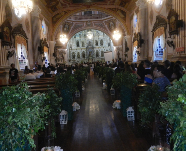 Eine Trauung in einer Kirche mit Gästen auf Bänken und im Gang sitzend und stehend, dekoriert mit Zimmerpflanzen, Skulpturen an den Wänden, Vorhängen an den Fenstern und hängenden Kronleuchtern, mit einem Bildschirm im Hintergrund.