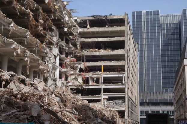 Ein große Abriss in einer Stadt, umgeben von intakten Gebäuden, Straßeninfrastruktur, Fahrzeugen, Bäumen und einem klaren Himmel.