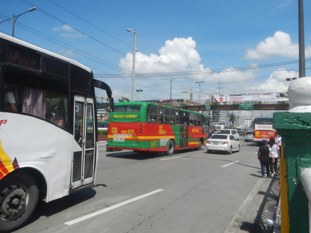 Eine Straßenansicht mit einem Doppeldeckerbus, anderen Fahrzeugen, Fußgängern auf dem Gehweg, einer Wand auf der rechten Seite, Laternen, Strommasten mit Drähten, einer Brücke, Bäumen und einer bewölkten Himmel im Hintergrund.