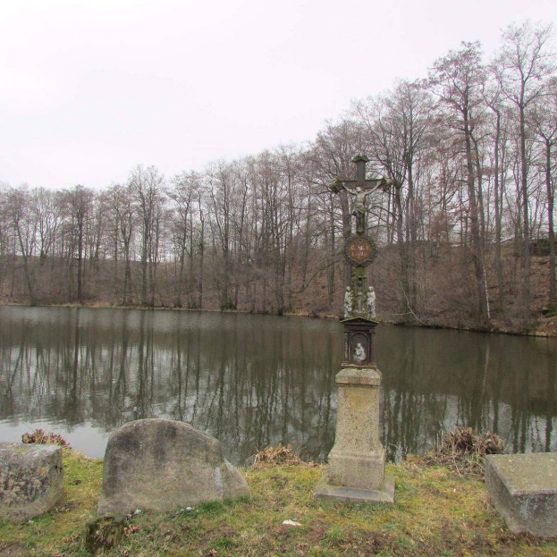 Ein Friedhof mit einem Kreuz in der Mitte eines Sees, umgeben von Bäumen und Grabsteinen, vor einem klaren Himmel.