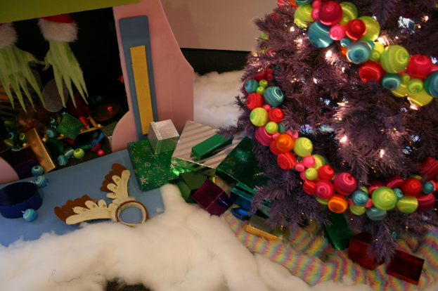 Eine Indoor-Szene mit einer Wand im Hintergrund, einem Baumwoll- und Wolltuch unten, einem geschm├╝ckten Weihnachtsbaum rechts, Geschenkboxen und Deko-Artikeln in der Mitte und einer H├╝tte mit Deko und Baumwolle oben links.