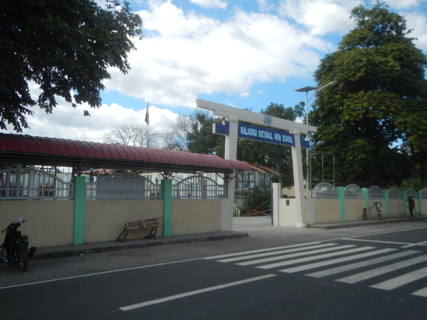 Einfahrt zu einer Schule mit einem vor ihr geparkten Motorrad, einer Straße mit weißen Linien und Zebrastreifen, einer Mauer mit einem Schuppen, einem Schild, Laternen, Bäumen, einem Fahnenmast und einem bewölkten Himmel.